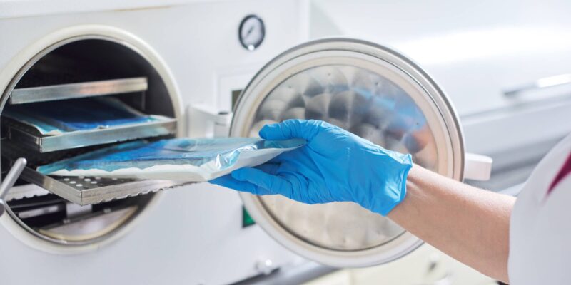 Mensch mit medizinischem Handschuh entnimmt verpackte Instrumente aus einem Sterilisationsgerät.